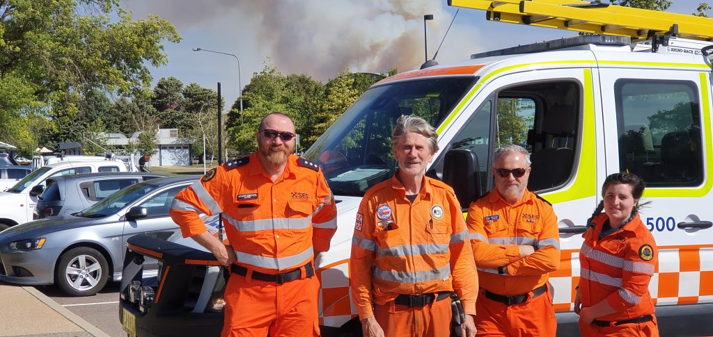 Four SES personnel standing next to an SES van. Smoke in the background
