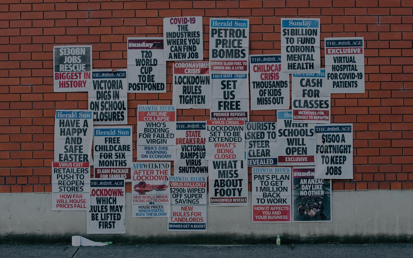 Multiple news posters on a brick wall. Headlines include 'Victoria digs in on school closures', 'What I miss about footy', 'PM's plan to get 1 million back to work' and 'Set us free'. 