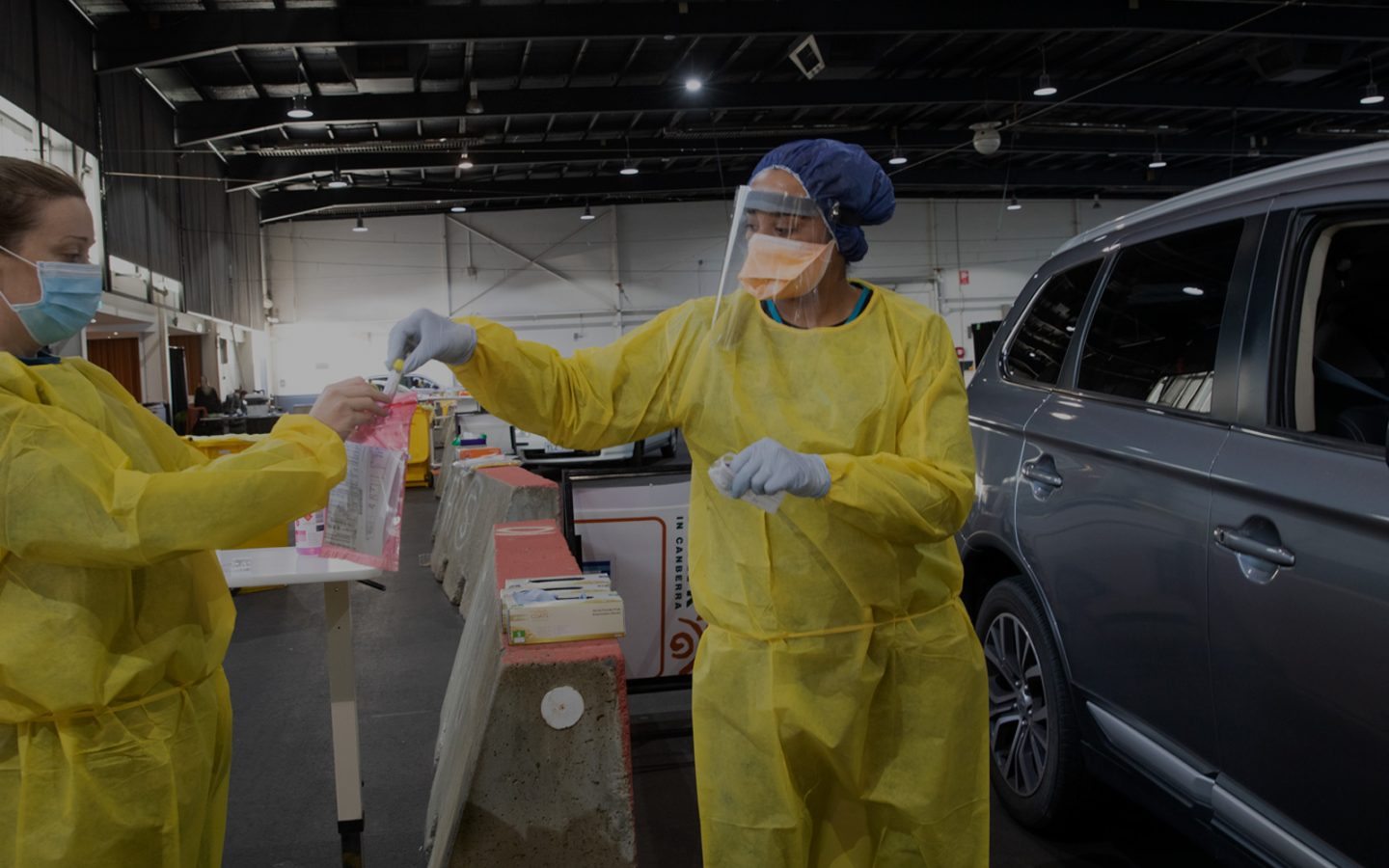 Two women wearing yellow protective gowns and face masks put a sample into a plastic bag. They stand beside a car in a large, indoor hall.