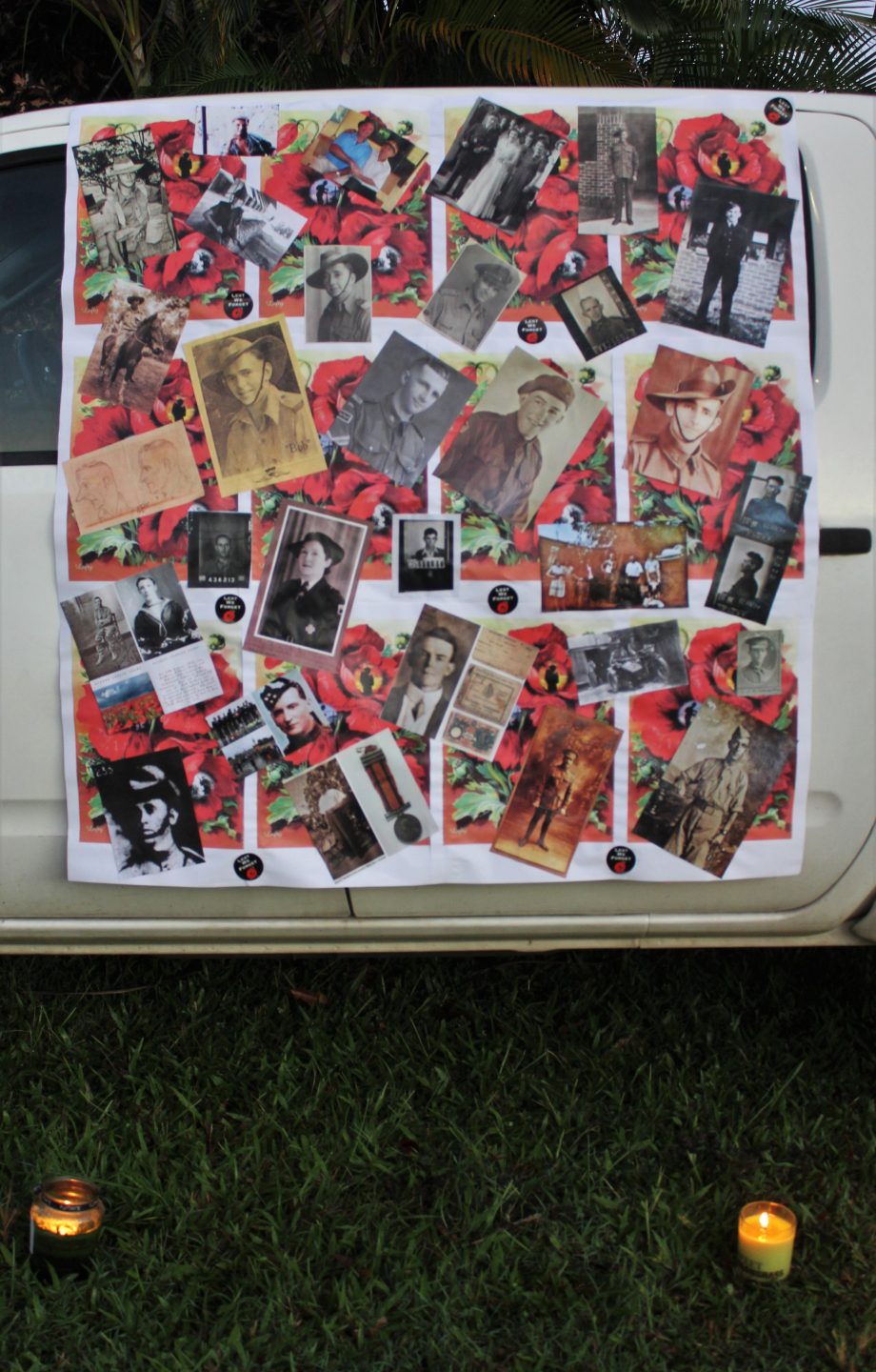 Collage of people dressed in army uniforms attached to side of white vehicle. Two candles on grass in foreground.