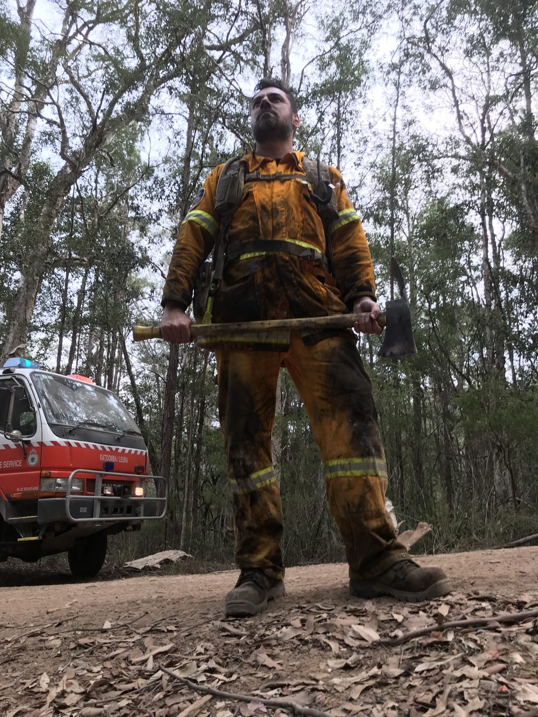 Firefighter holding axe covered in soot on a dirt road with trees in the background. 