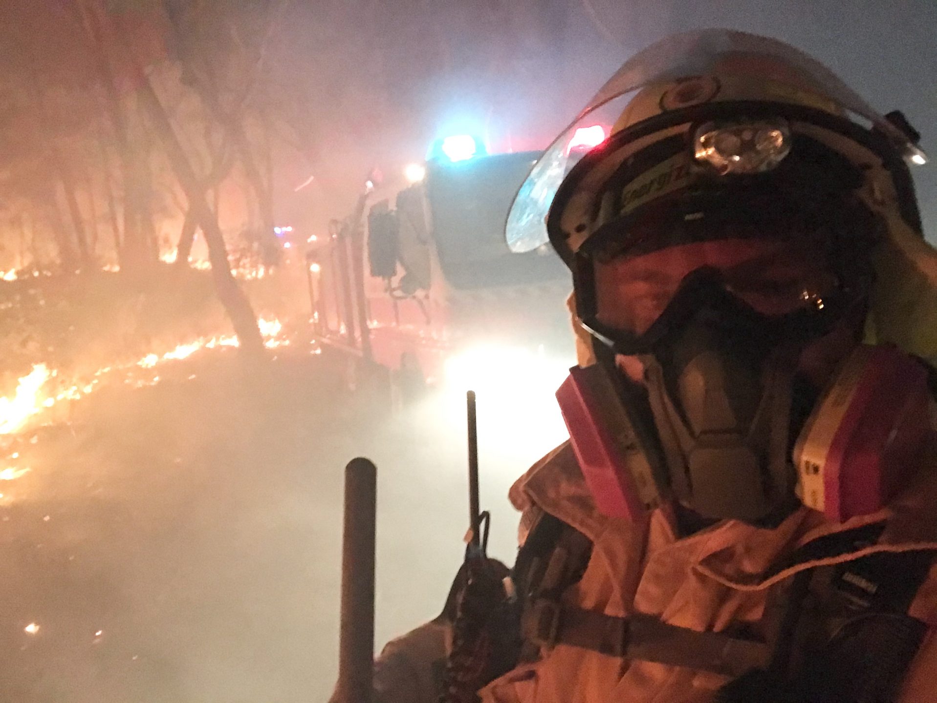 Fireman wearing a mask and googles surrounded by smoke standing in front of a truck and bush land that's on fire.