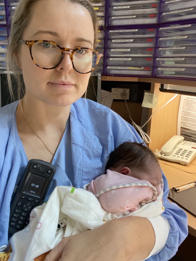 An obstetrics doctor in blue scrubs, holding a newly delivered baby who is swaddled in a white blanket and pink suit, in a medical ward office