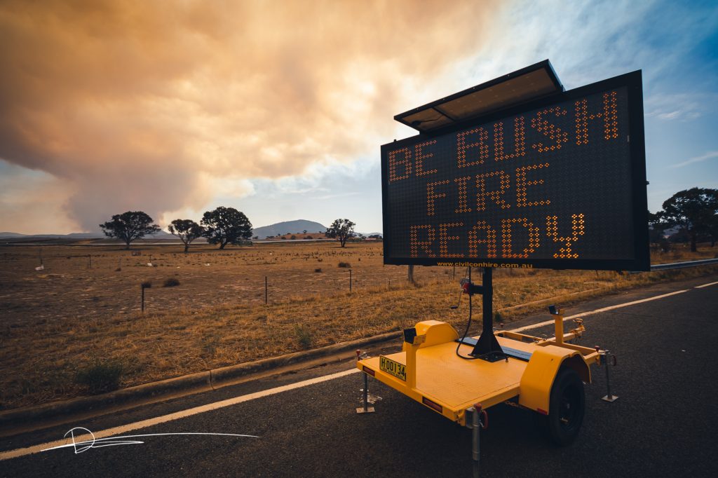 Photo shows smoke from a fire in the background, a dry paddock and a sign saying be Bushfire Ready.