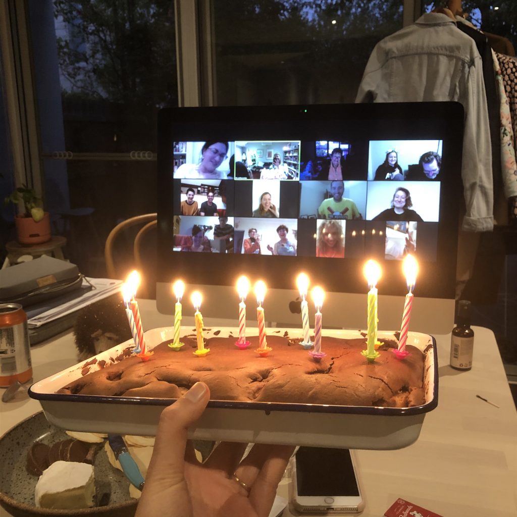 Birthday cake with ten lit candles, being held in front of a computer screen with 16 people in squares (Zoom). 