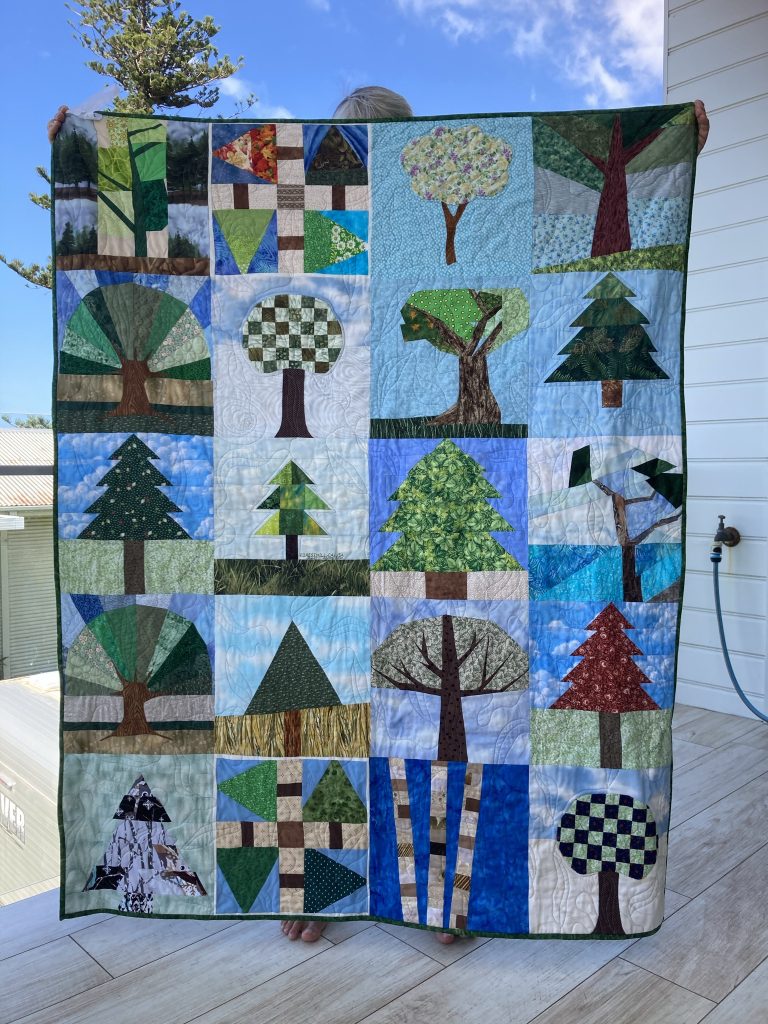 A full quilt being held up. Each panel shows a different tree in a different season. Colours are green, blue, red, black and white. 