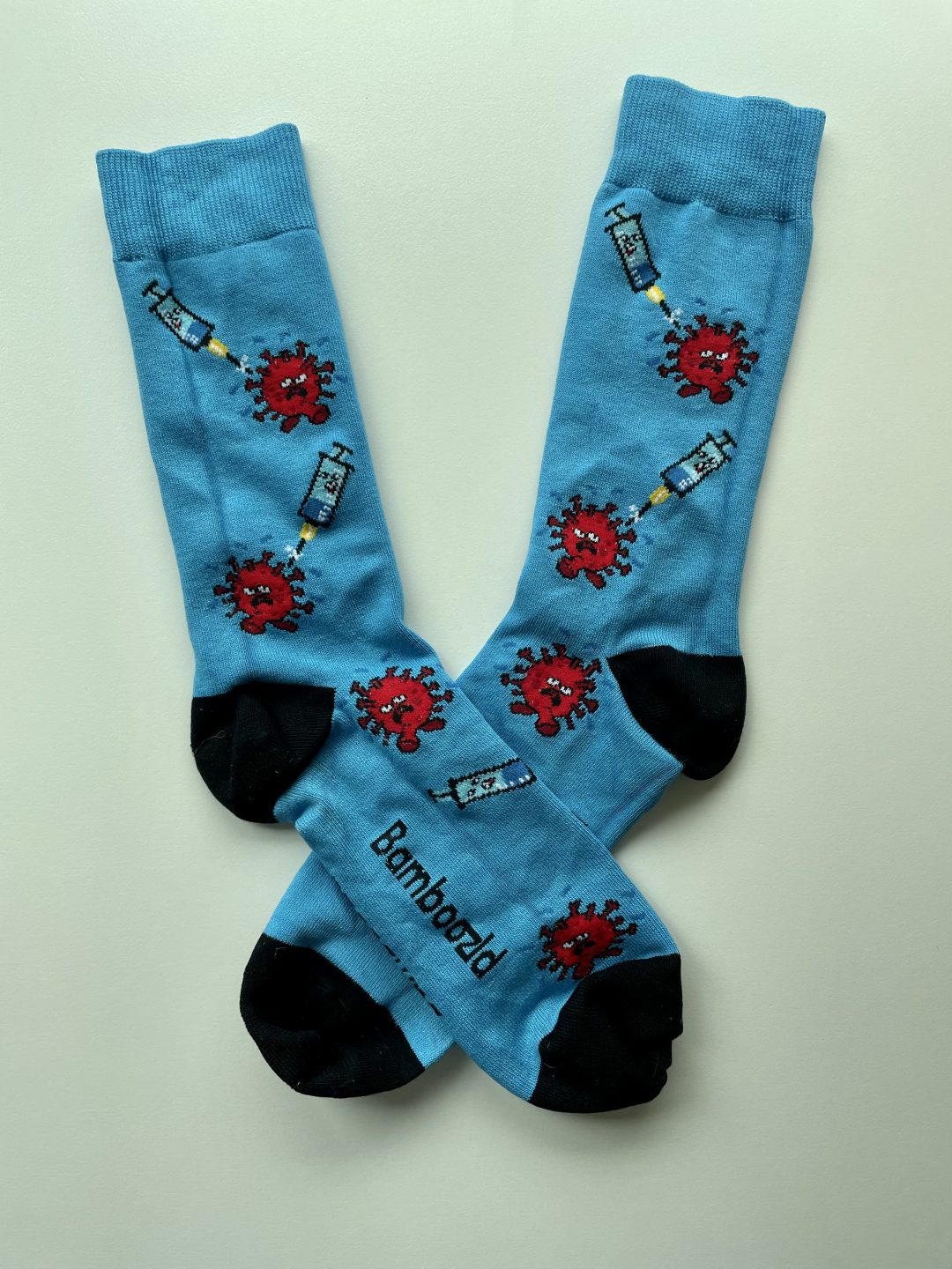 Two socks. The left is placed over the right. Both are bright blue, with black toes and heels, with repeating decoration of a red virus and a blue syringe facing towards it. 