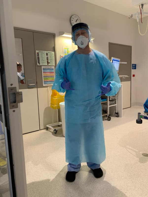 A man standing in a hospital setting wearing a full suit of blue personal protective equipment including a head covering, mask, long blue outerwear, gloves.  