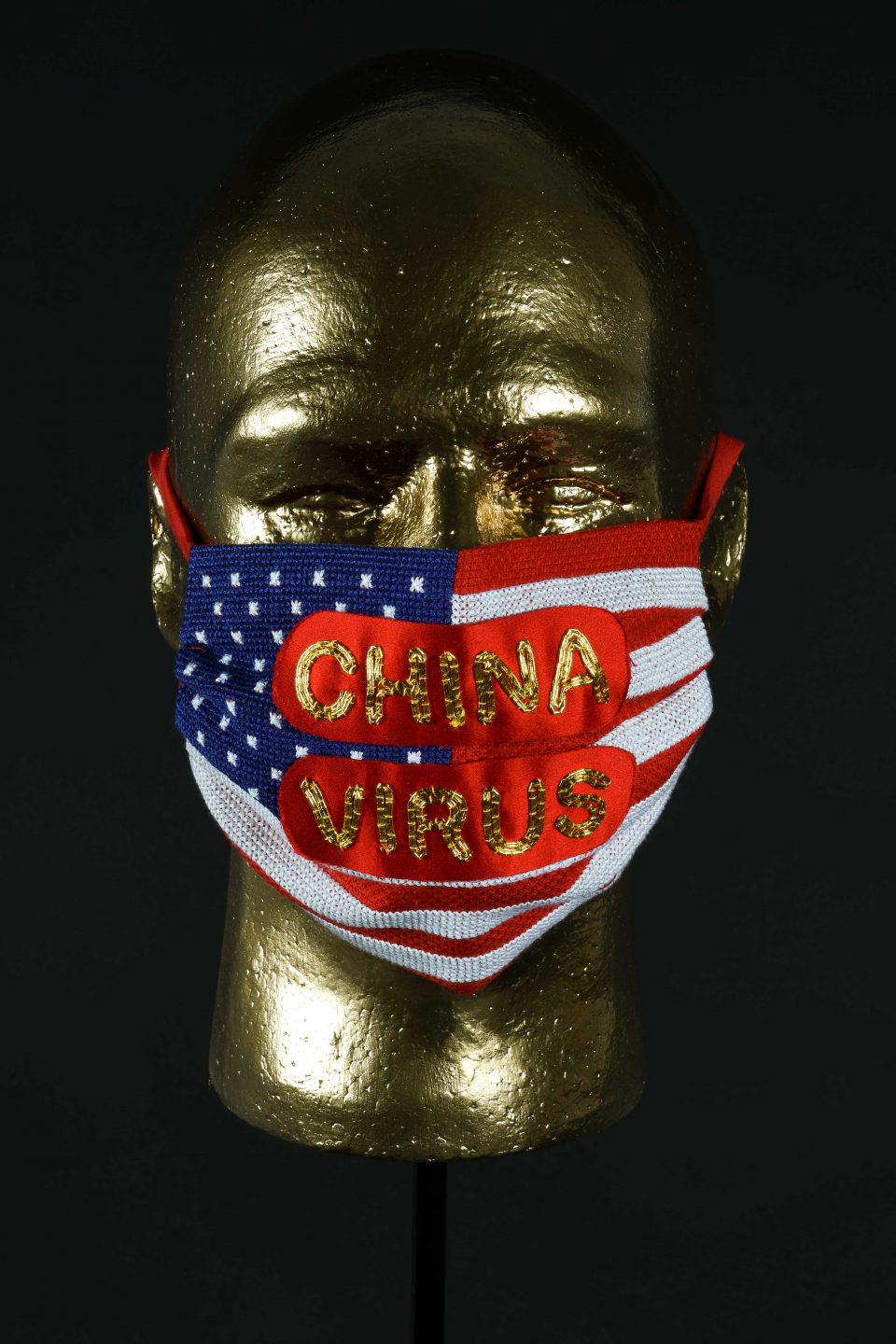 Golden mannequin head with a handstiched mask featuring the American flag overlaid with red satin with China Virus stitched on top. 