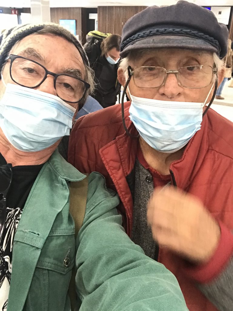 Two caucasian men, both wearing blue surgical masks. A selfie. Both are wearing hats. One has black glasses and a green jacket. The other silver glasses and a red jacket.
