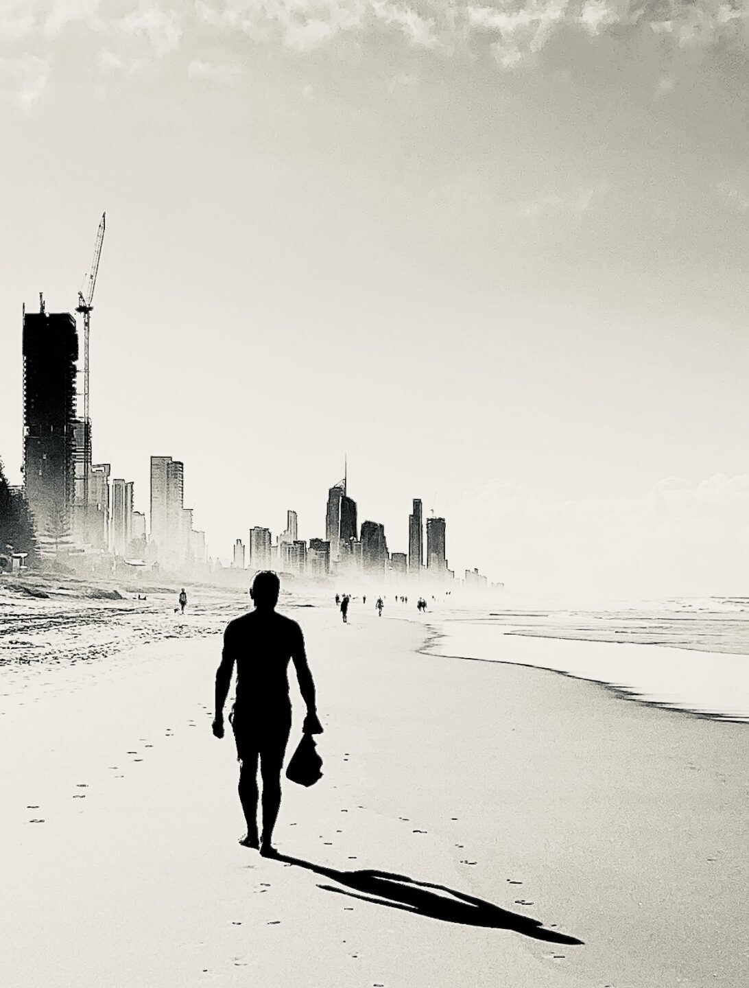a silhouette of a man walking on a beach with tall buildings to his left. 