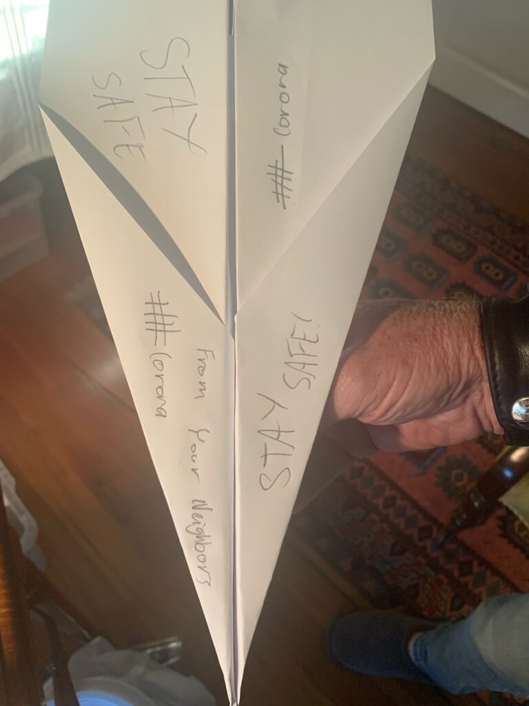 One white paper airplane with 'stay safe' and #Corona written on it in pencil. It is being held my a hand, and a polished wooden floorboard with a rug are visible. 