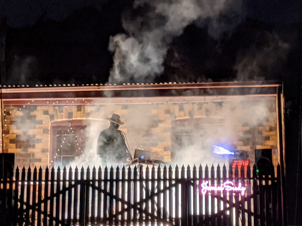 Nighttime. A brick building partially concealed by smoke, with a male hatted figure silhouetted. In the foreground is the profile of a picket fence. 