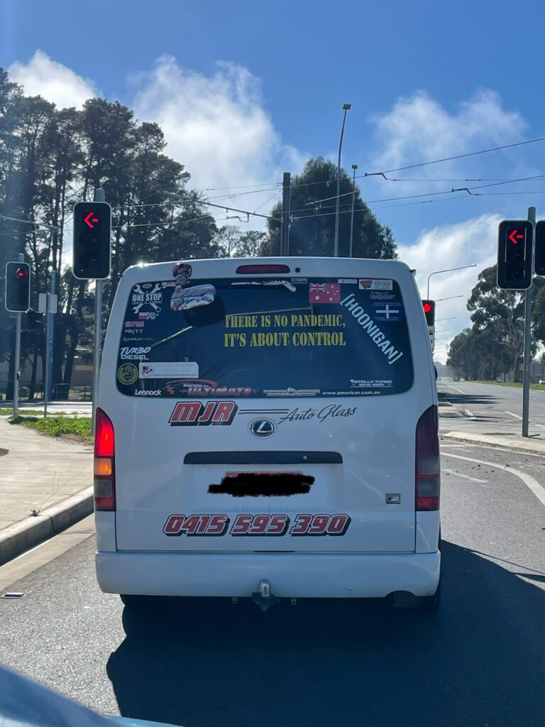 Rear of a white van stopped at lights in daytime. A vinyl sticker on the back window reads THERE IS NO PANDEMIC IT'S ALL ABOUT CONTROL. The window has a lot of other stickers. 