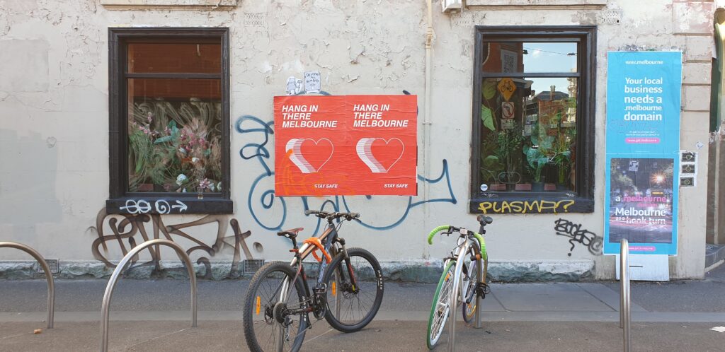 A photo of the side of a Melbourne building with two red posters on it. four bike racks and two bikes are in the foreground. 