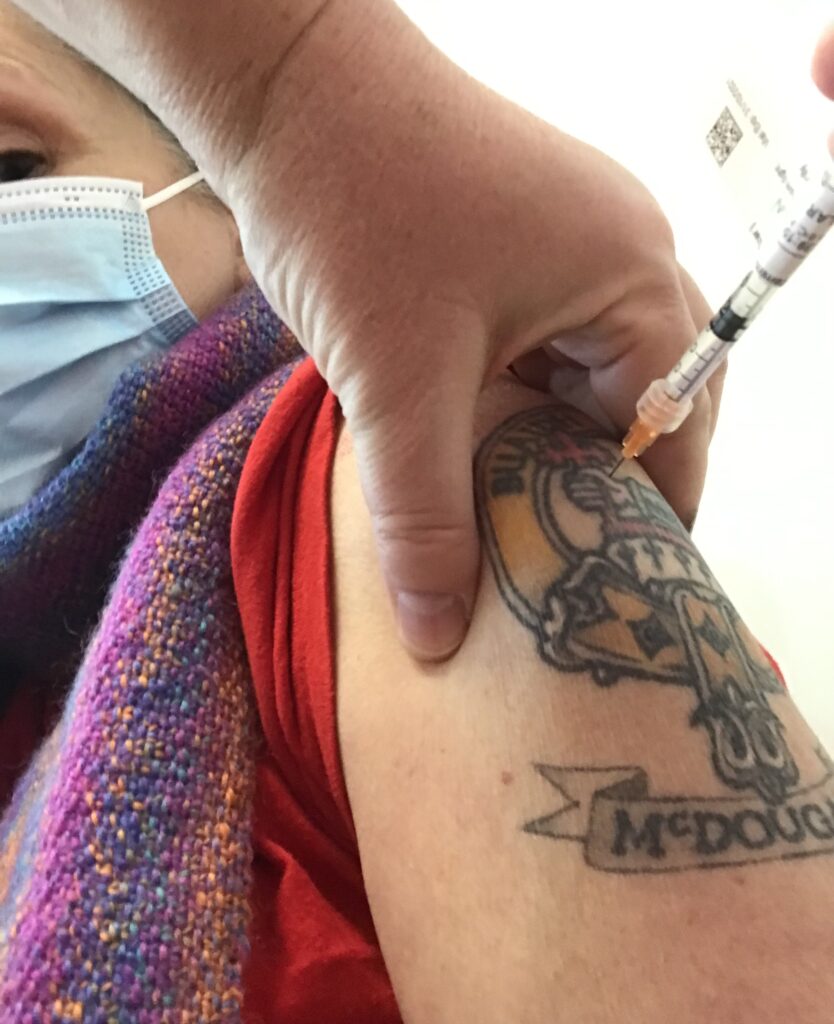 A close up of a covid-19 vaccine needle in a person arm. The person is white and has a tattoo on their arm.