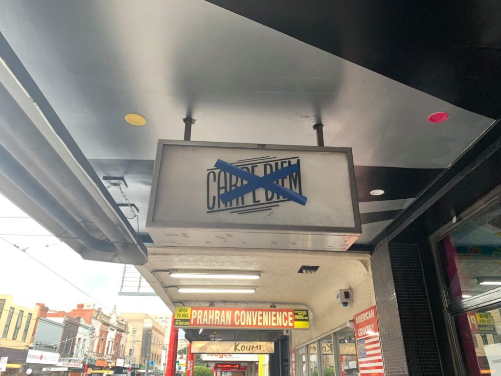 A light box sign bolted to a shop veranda with blue tape crossing out the name of the venue.