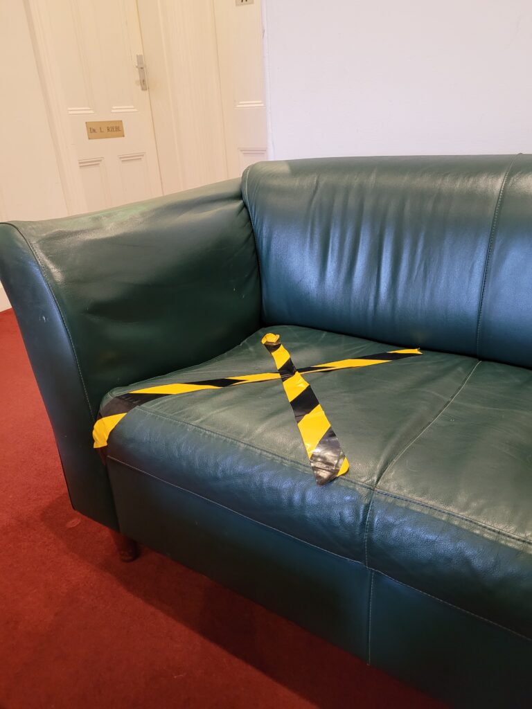 A forest green couch with black and yellow tape in the shape of an X to mark where a visitor can not sit.