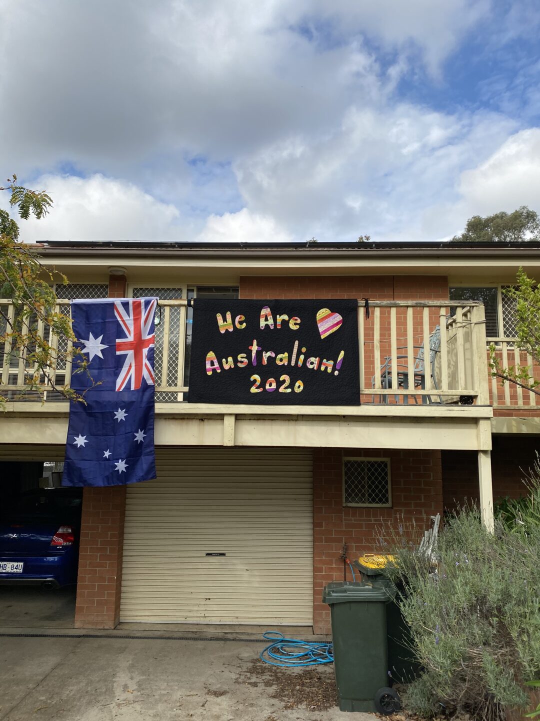 a handmade quilt with the words 'we are Australian 2020' suspended on a veranda next to an Australian flag. 