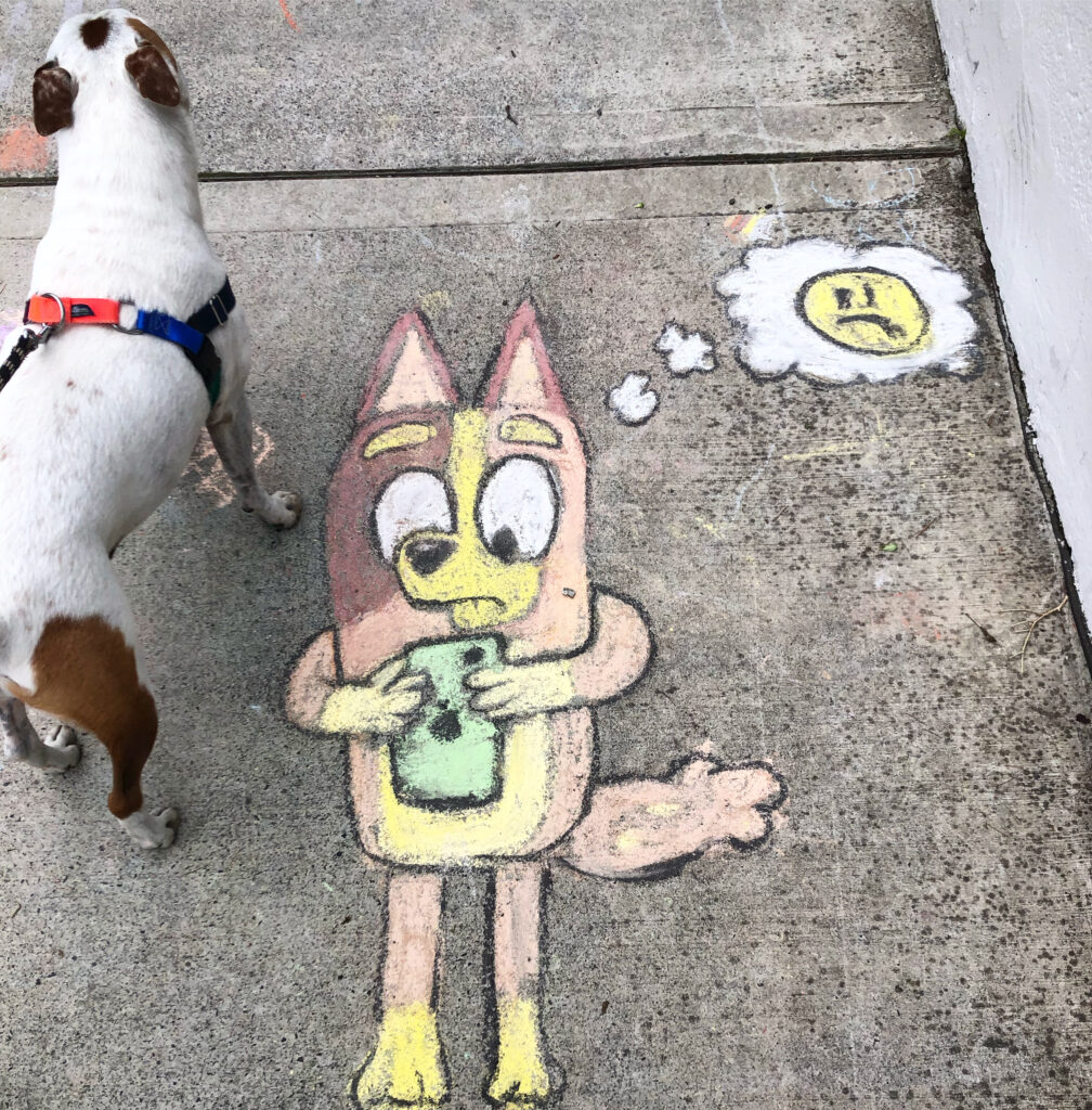 A yellow, brown , white and blue chalk drawing with black detailing. It is a chalk drawing of the character Bingo from Blue, looking at a green mobile phone woth a worried expression. A thought bubble contains a yellow emoji-like sad face. In the top roght of the photo is a small dog, probbly a jack russell, white with brown spots. The dog has a red and blue harness. 