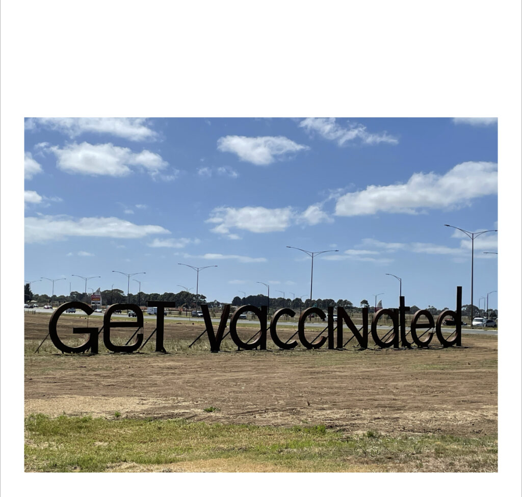 Daytime scene, with grassy expanse and trees, a road and street lamps in the disance. The blue sky has several wispy white clouds. Large black 3D letters read 'GET VACCINATED'