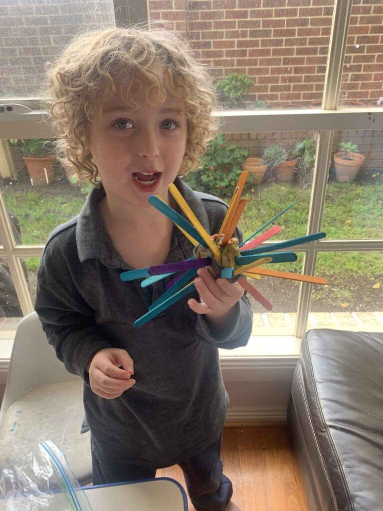 a young boy with curly blond hair holding a sculpture of a coronavirus spore made from pop sticks. 