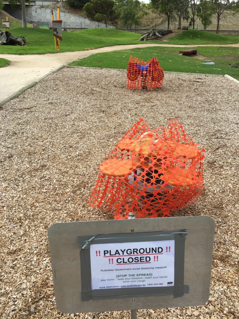 A playground in a public park with equipment wrapped in orange barrier mesh and a sign that says 'playground closed'