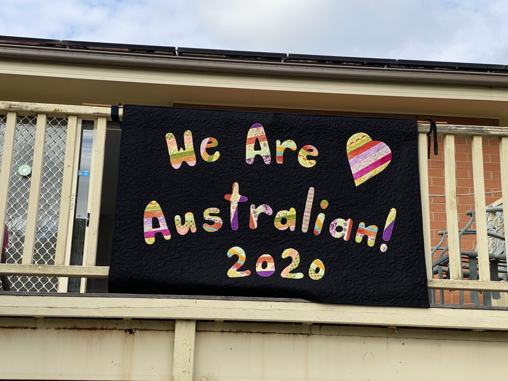 a handmade quilt with the words 'we are Australian 2020' stitched on it. The quilt is suspended from the side of a veranda balcony.