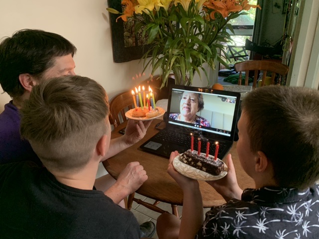 Children looking at a computer screen holding cake while talking on Zoom with their great grandmother