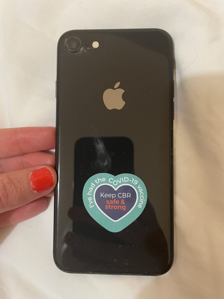 Black apple phone with a blue and purple heart sticker at the bottom, reading 'I've had the COVID-19 vaccine / Keep CBR safe & strong'