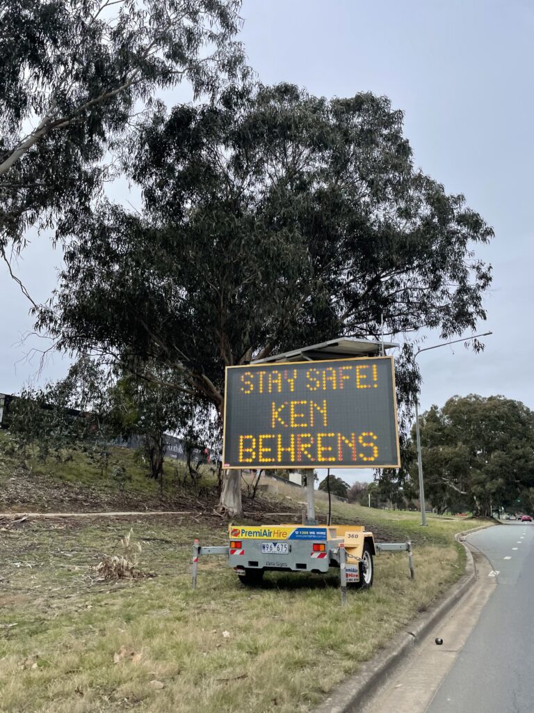 Overcast daytime scene. An electric sign by a road reading STAY SAFE KEN BEHRENS in orange illuminated lettering. Behind the sun is a large eucalyptus tree. 