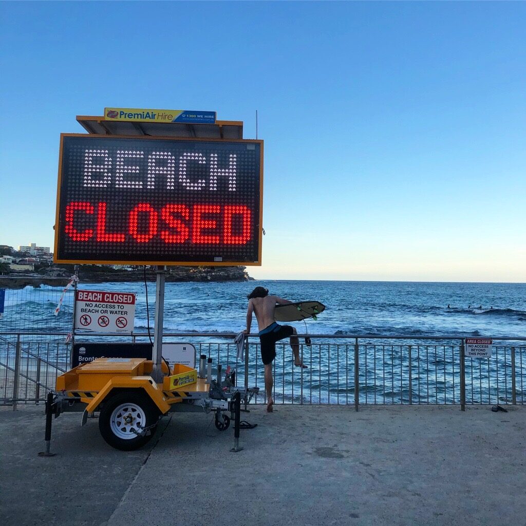 A beach scene, viewed from a concrete pavilion. An electric sign reads BEACH CLOSED in white and red lettering. Next to this, a shirtless young man carrying a surfboard is stepping over the railings. In front of him is the ocean, and in the distance coastal properties are visible. It is probably in the early morning. 