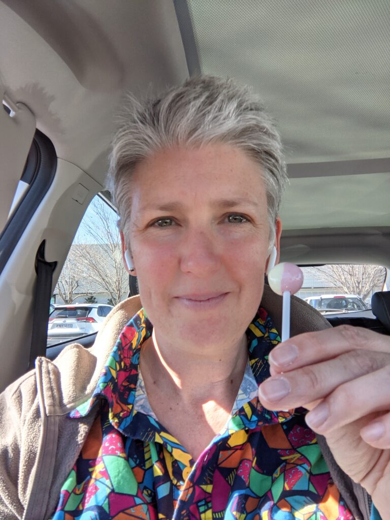 A white person with grey hair sitting in a car holding a pink lollipop.