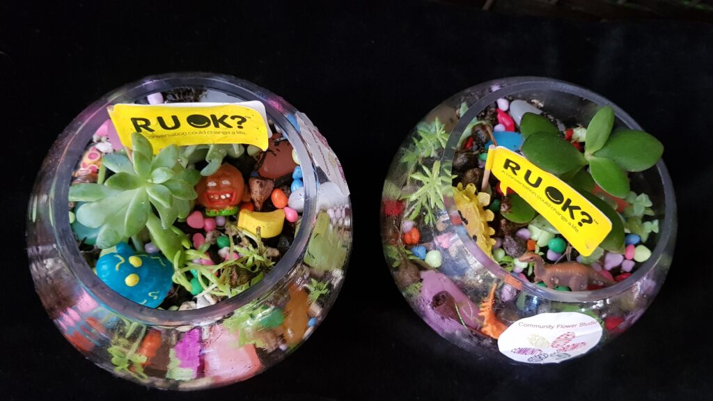 Two terrariums with small yellow flags in them that say R U OK ? 
