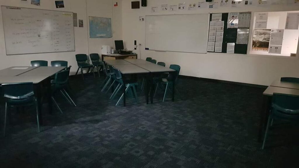 A classroom with white walls whiteboards with text written on it, and dark blue-grey carpet. Two rectangular tables with chairs are placed in the corner, and there are posters on the walls. The room is empty. 