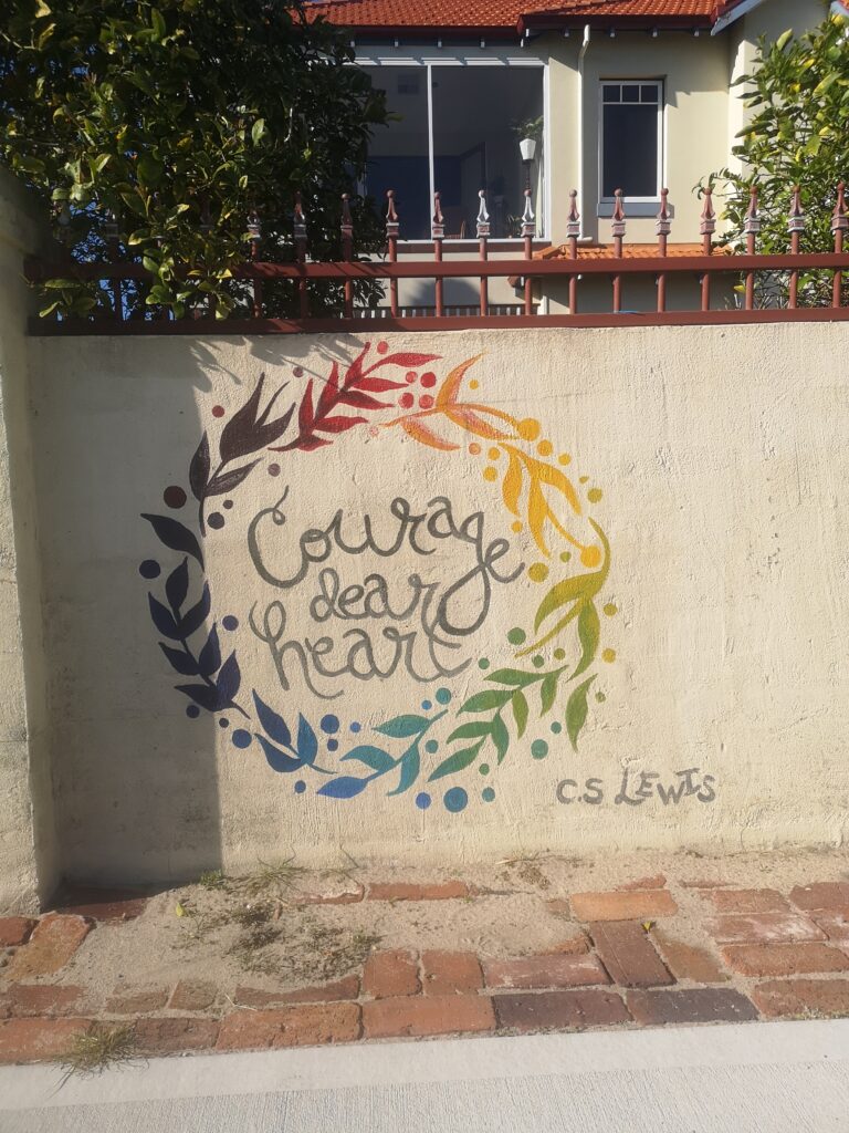 A daytime scene, with a concrete wall in front of a residential property. On the wall is stencilled in rainbow colours a wreath, with cursive text within reading 'Courage dear Heart' and an attribution to C.S. Lewis in the bottom right. 