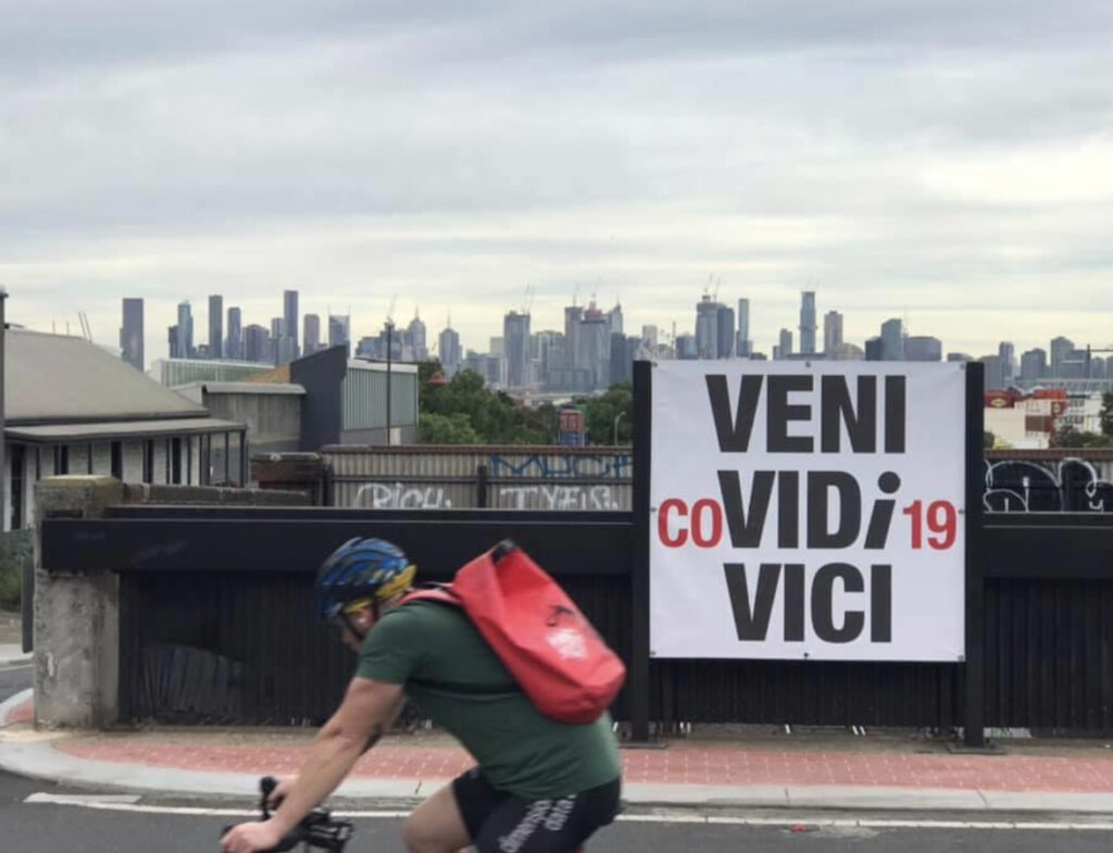 a view of the melbourne skyline behind a poster that says 'veni COvidi19 civi'. There is a bike rider in a green t shirt and red backpack in the forground.