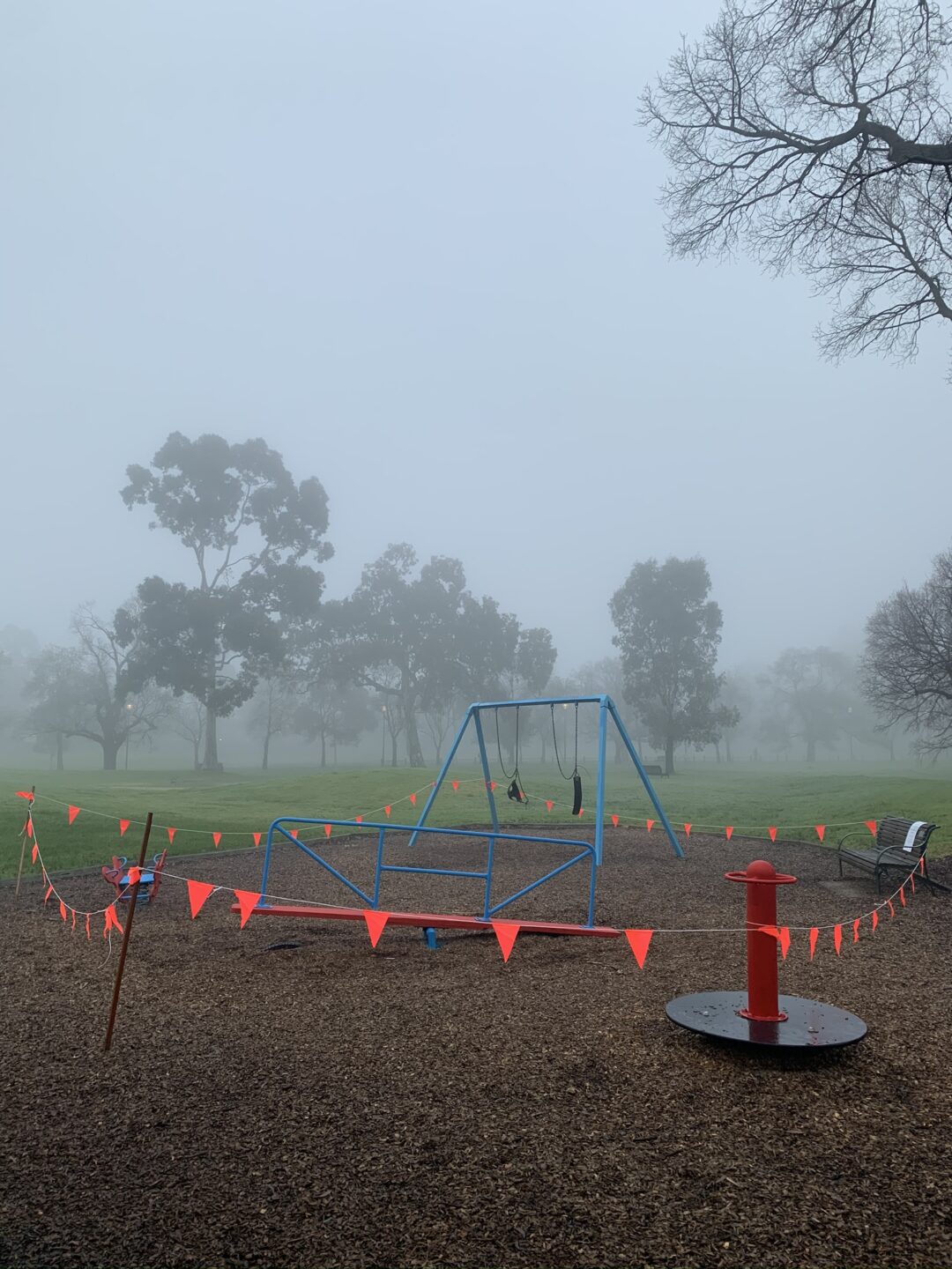 A frosty, foggy winter's morning. Blue children's play equipment is cordoned off by red and orange bunting.  
