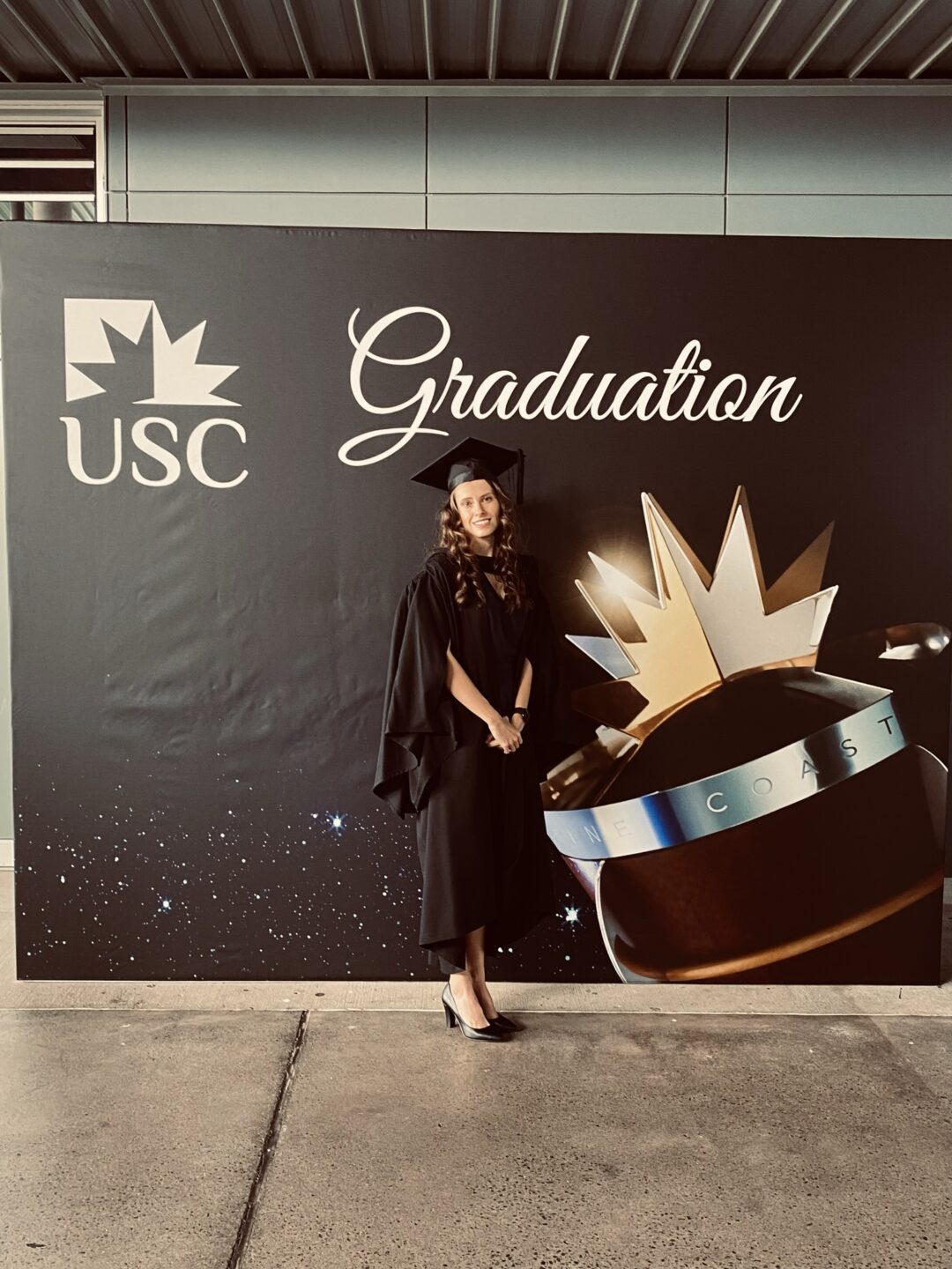 A women with golden brown hair in a black graduation robe stands in from of a sign that says 'Graduation'