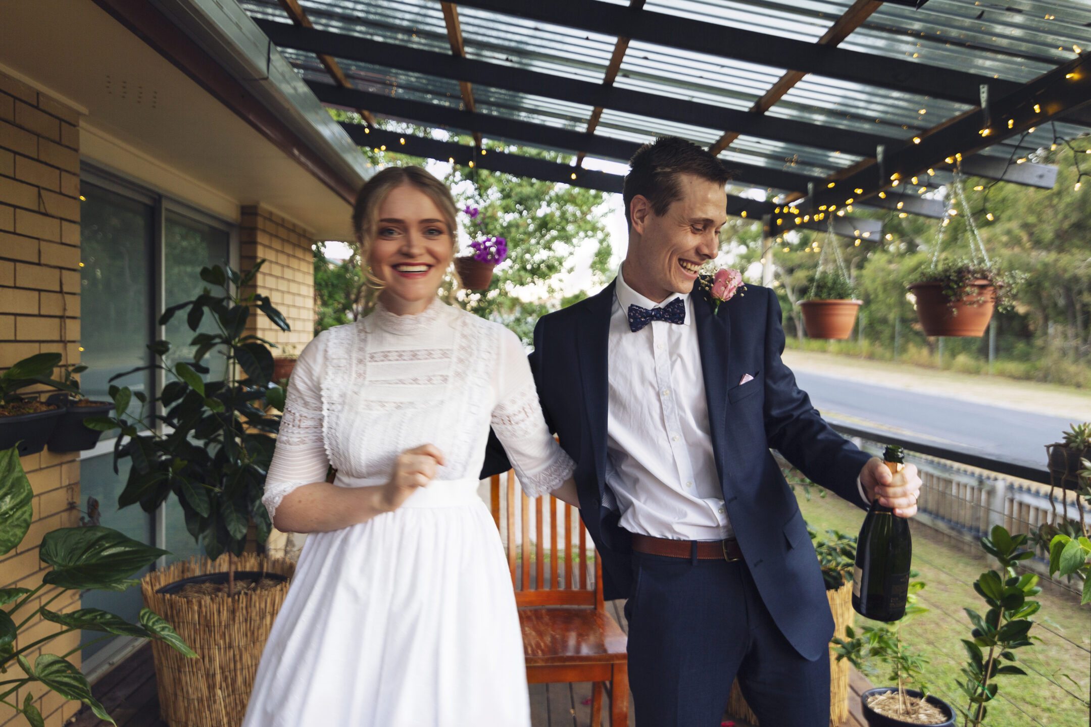 a young woman in a white long sleeved wedding dress and a young man in a navy suit and navy bow tie embracing and holding a bottle of champagne.