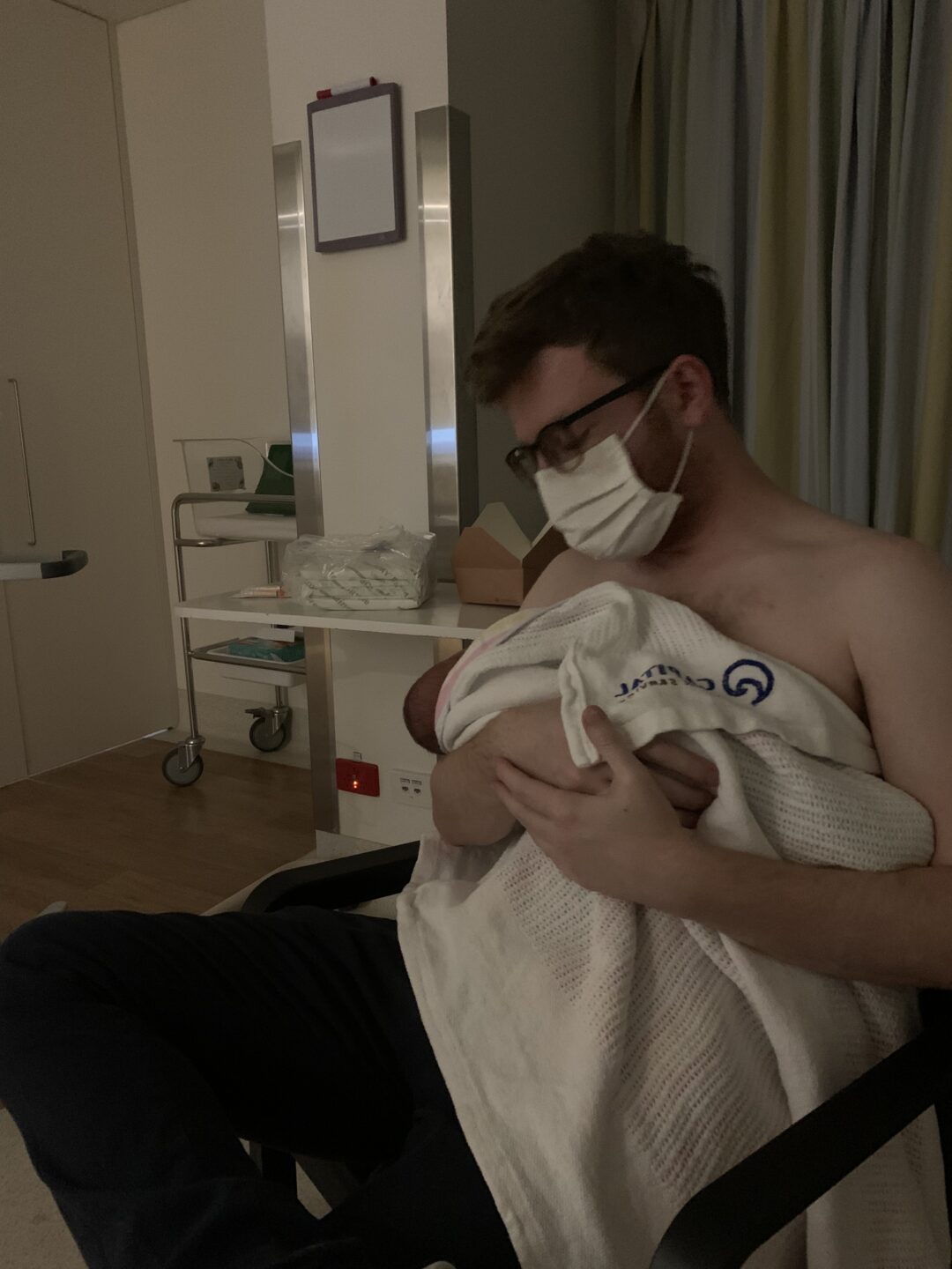 A caucasian man with brown hair in a white medial mask holding a baby wrapped in white blankets