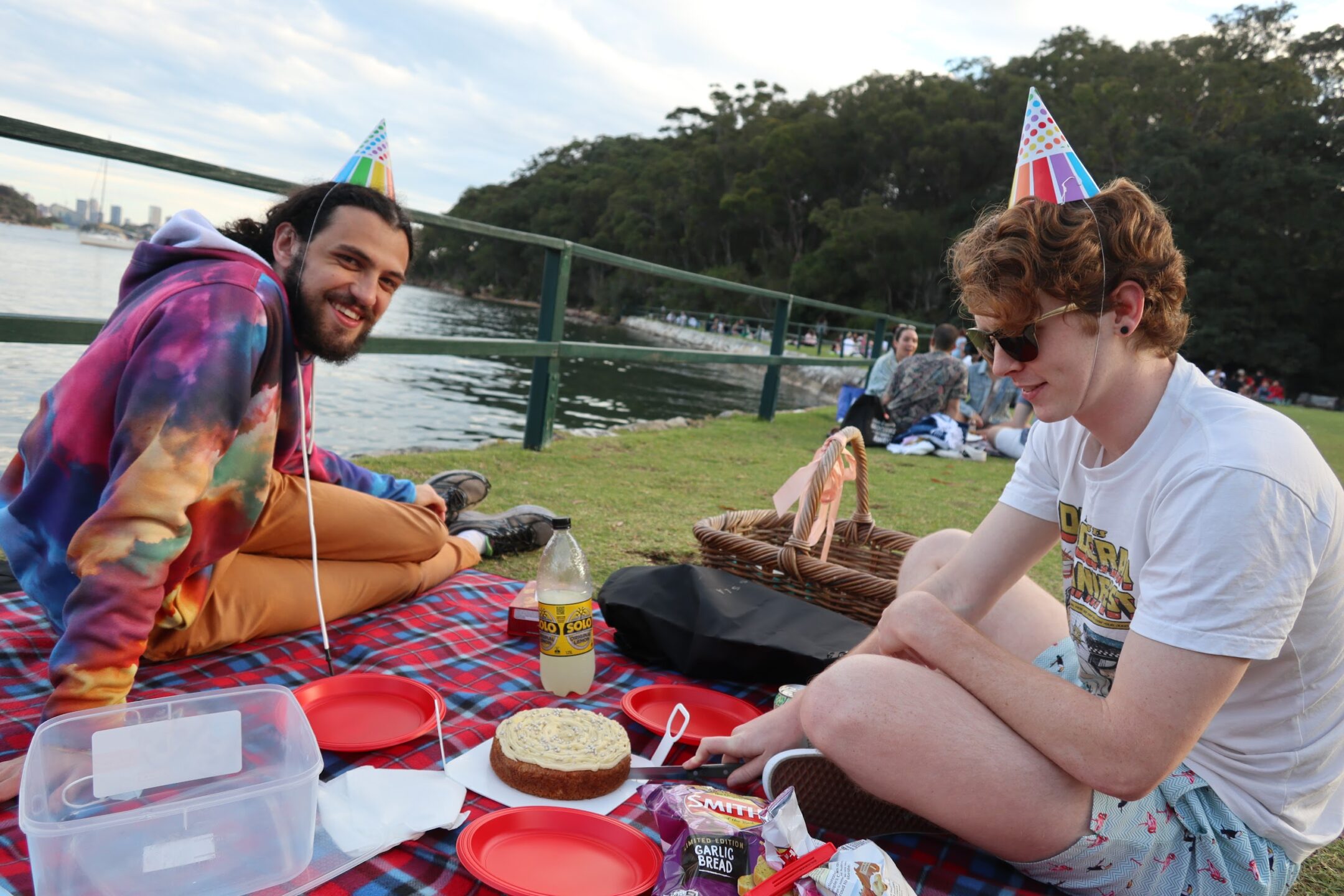 Two people in colourful party hats sitting on a picnic rug eating food