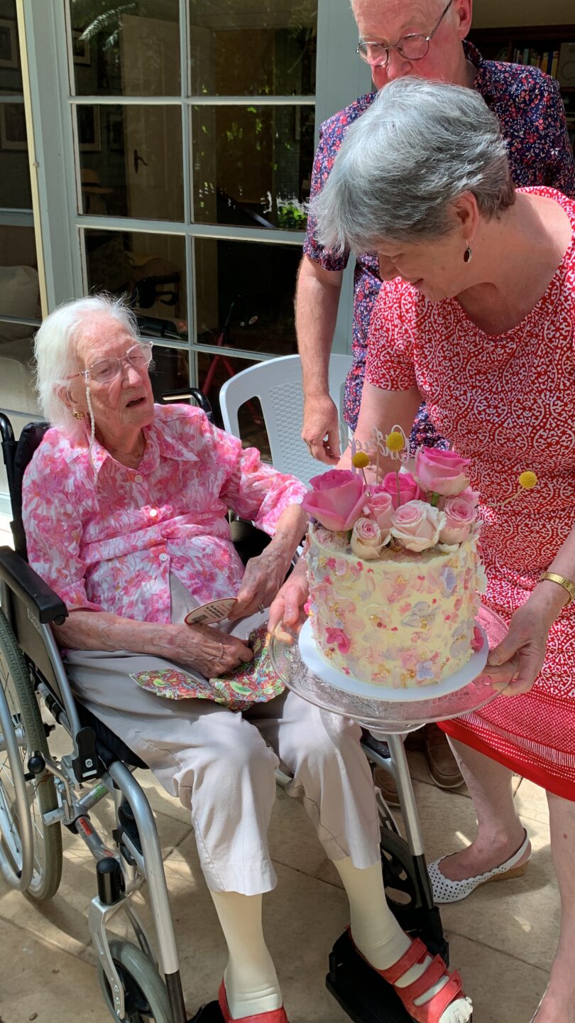 An elderly Caucasian woman sitting in a wheelchair receiving a birthday cake.