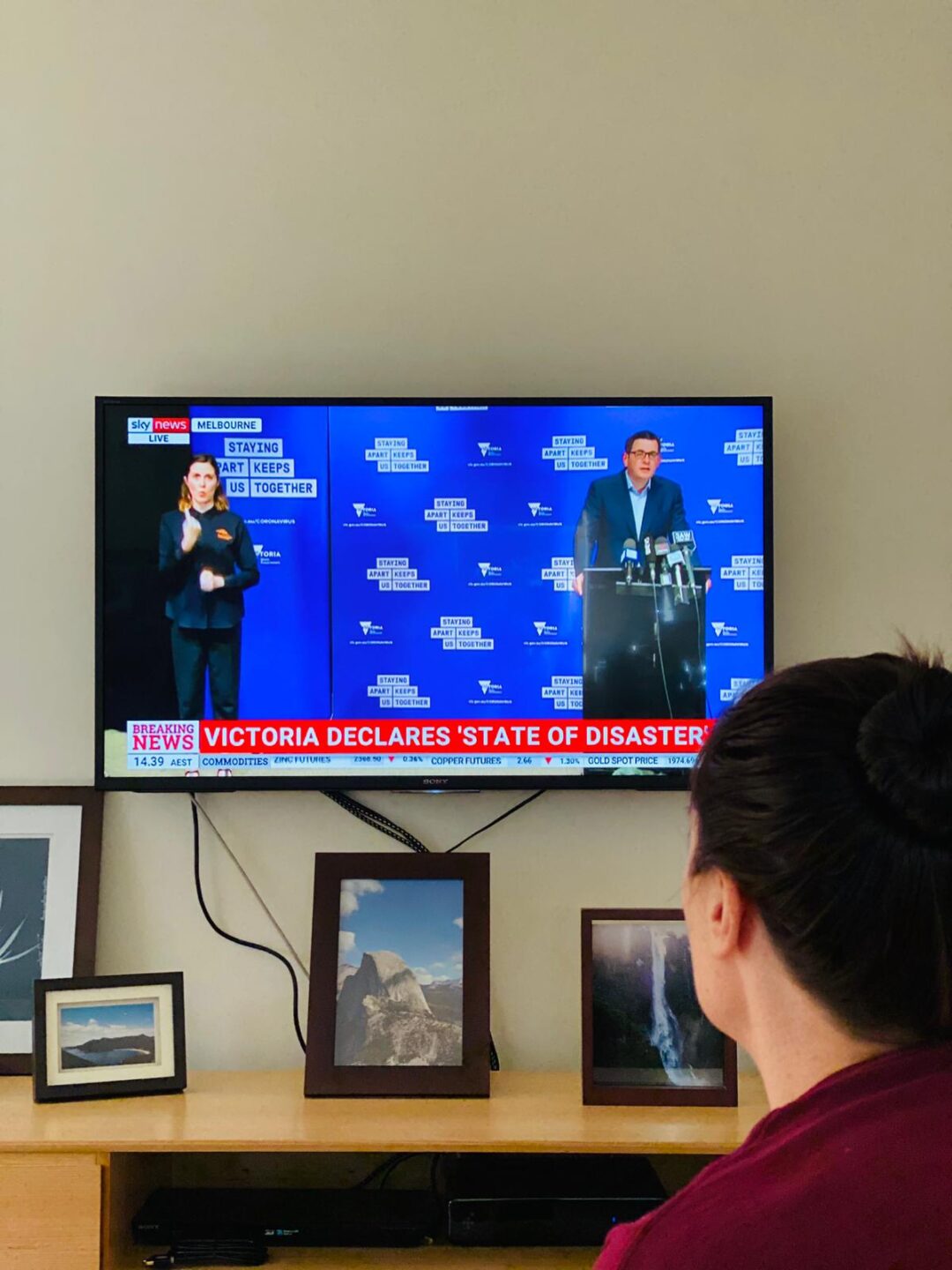 Woman watching TV displaying Daniel Andrews at a press conference. Text reads 'VICTORIA STATE OF DISASTER'. 