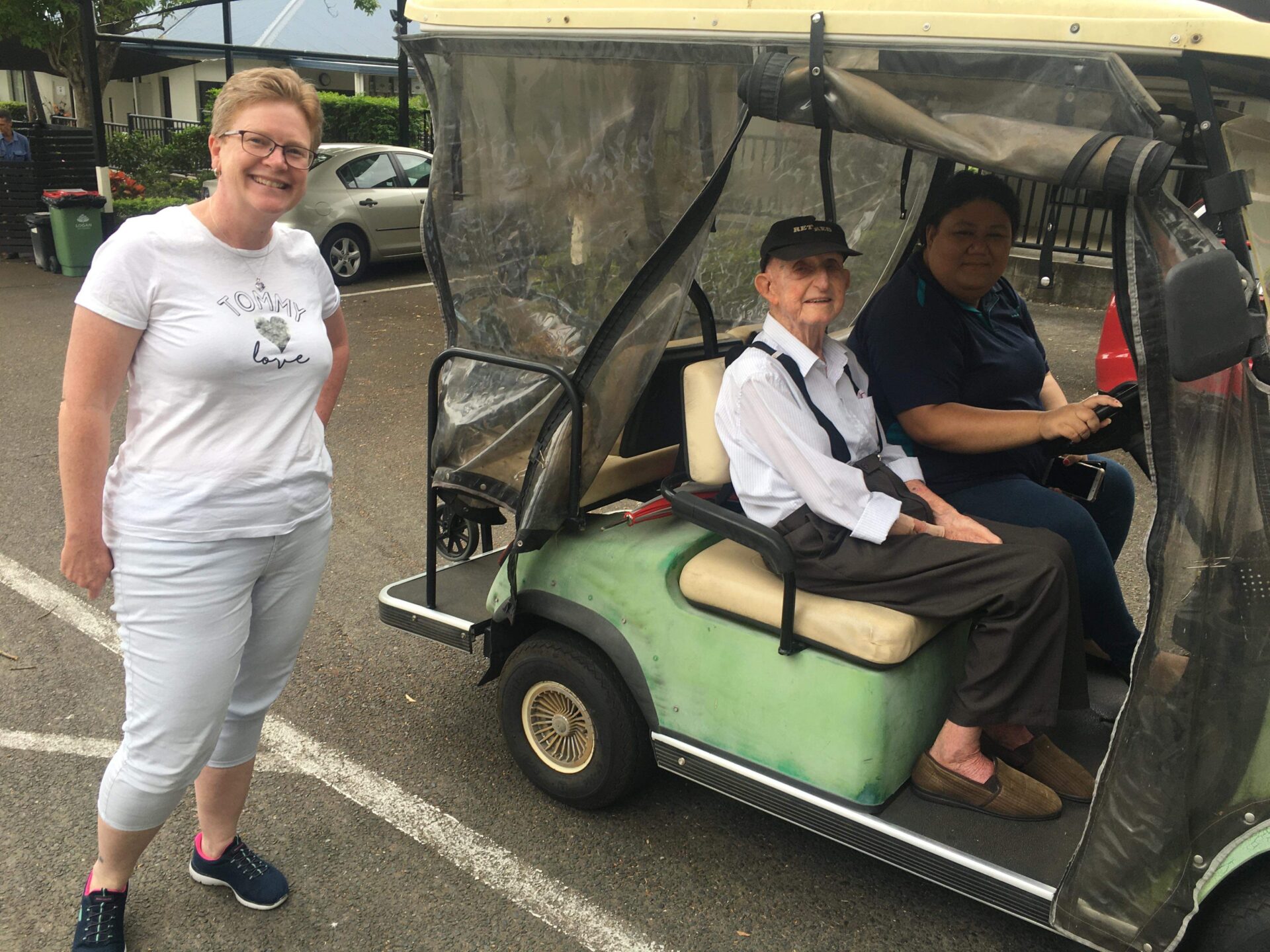 A Caucasian women with short blonde hair standing next to a golf cart which her grandfather, an elderly Caucasian man in a black cap sits alongside a nursing home worker.